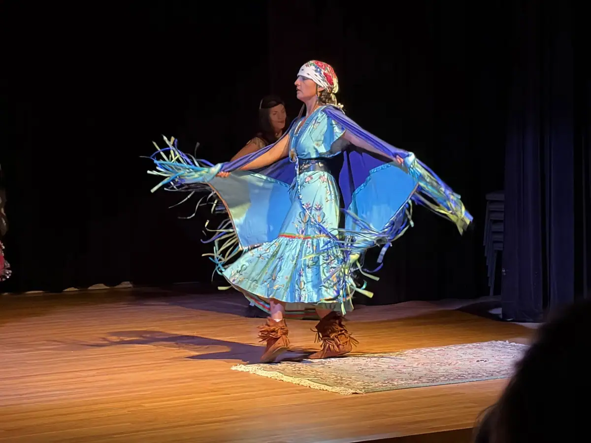 A dancer in a blue costume performs on a stage while holding wide, fringed fabric.
