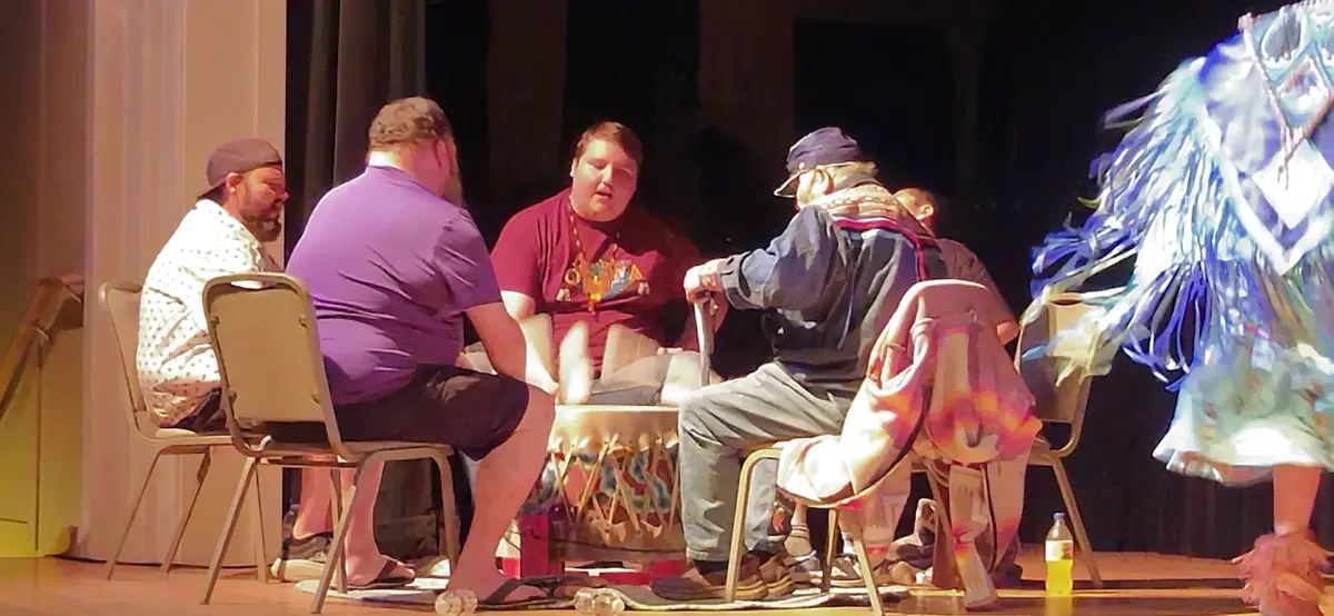 Four people sit around a decorated low table on a stage while a feathered-costume performer stands at right.