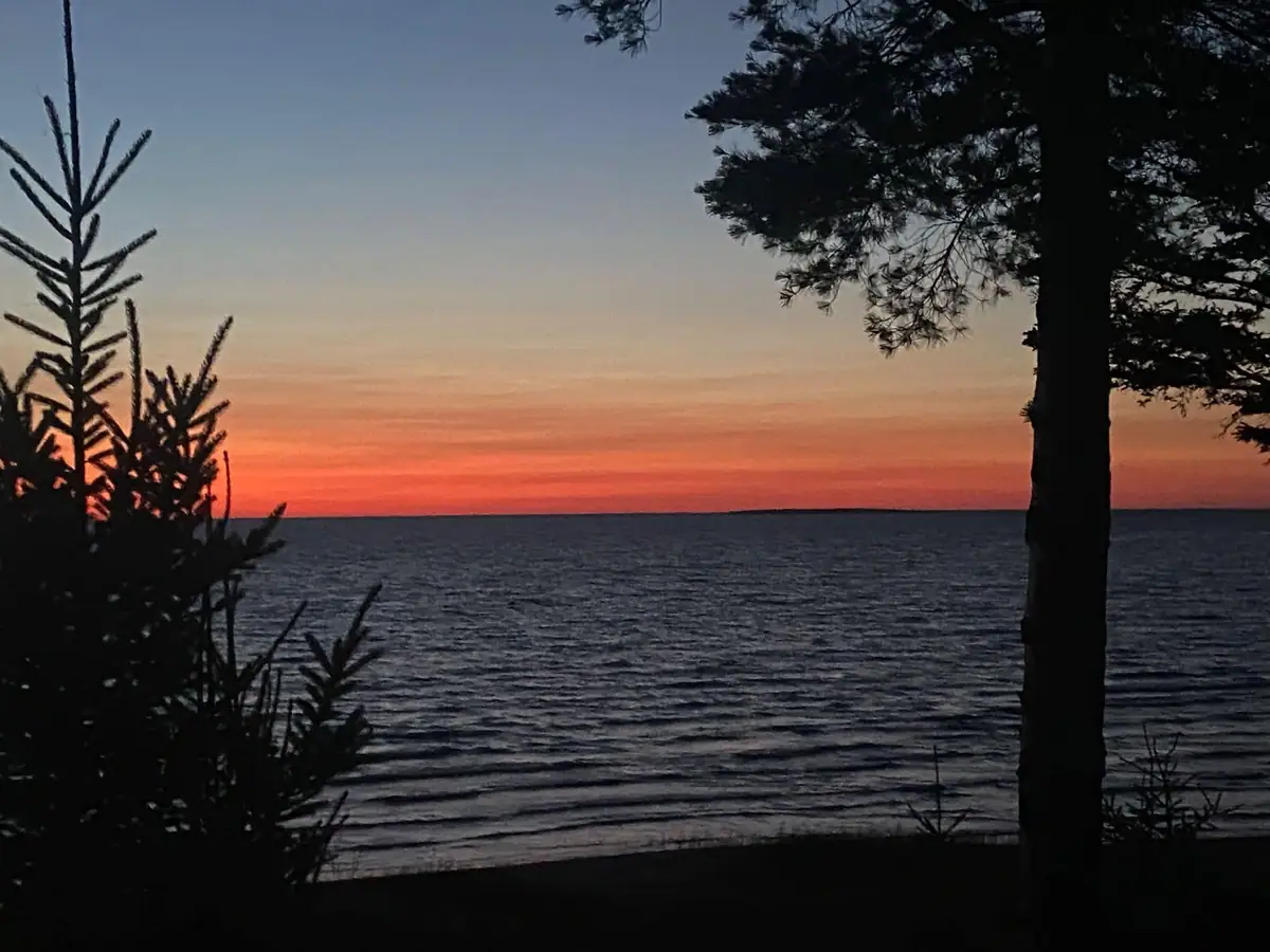 A red-orange sunset glows over rippling water, framed by dark evergreen trees.