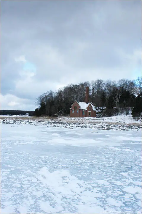 A red brick lighthouse-like building stands beyond a broad expanse of frozen water and snow.