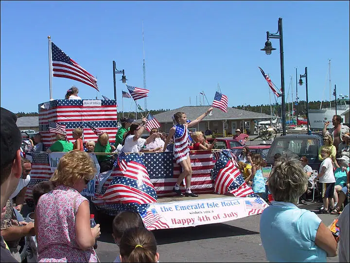 Patriotic parade float covered in American flags, with people gathered along a waterfront street.