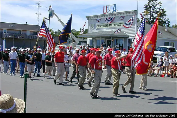 Marchers carrying flags pass a decorated storefront while spectators watch from the sidewalk.