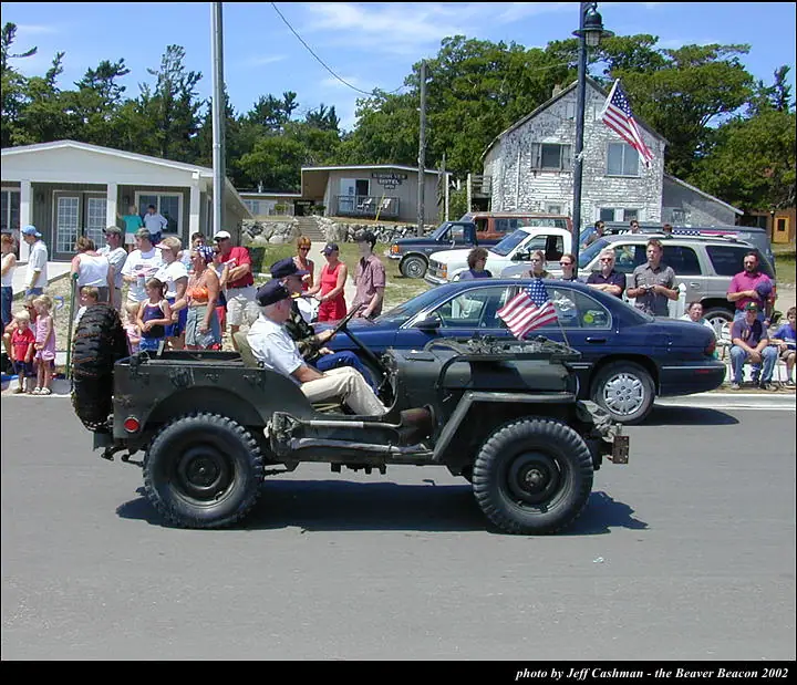 A dark green jeep drives past spectators during a parade, with flags, cars, and wood-frame buildings behind it.