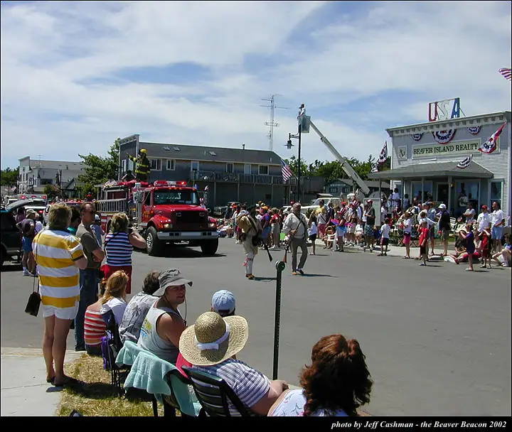 A red fire truck leads a street parade past a crowd gathered outside storefronts.