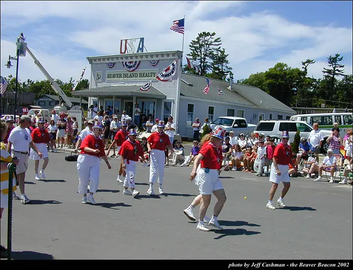 Marchers in red shirts and white trousers pass a decorated realty office while spectators line the street.