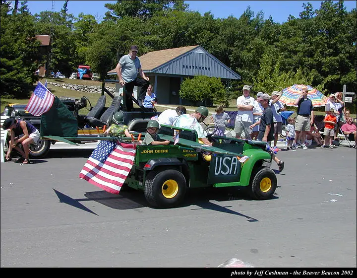 A flag-decorated green utility vehicle moves through a parade past spectators and colorful umbrellas.