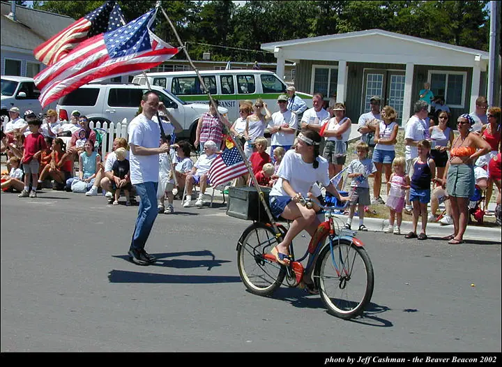 A cyclist carrying flags rides past spectators while a man walks alongside in a street parade.