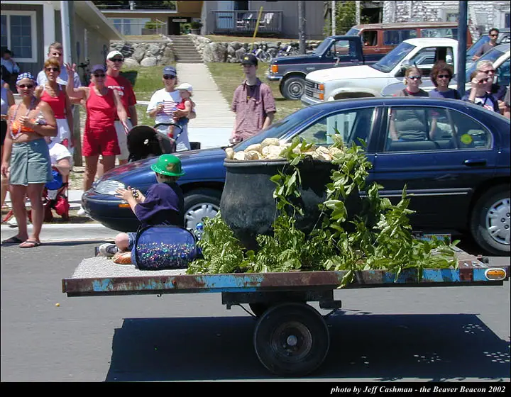 A leafy parade float with a large pot passes spectators and parked cars on a sunny street.