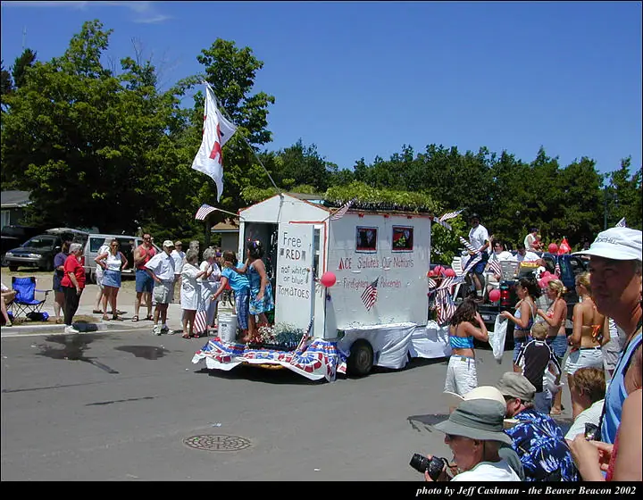 A decorated trailer float passes spectators, with signs honoring firefighters and policemen and offering tomatoes.