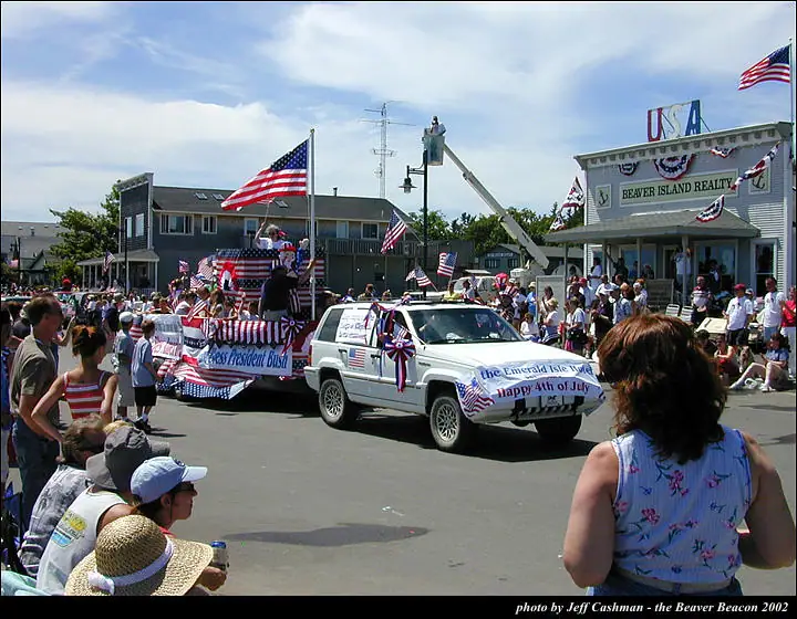 A patriotic parade passes a storefront with a decorated SUV, float, flags, and a large crowd.