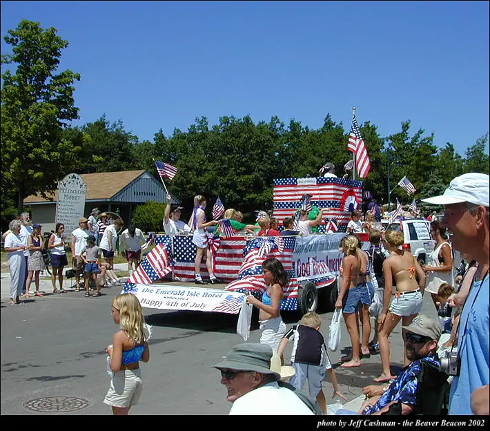 A patriotic parade float covered in American flags and bunting moves past spectators on a sunny street.