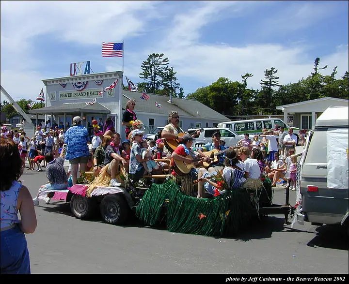 A decorated parade float with guitar players passes spectators on a street beside a commercial building.