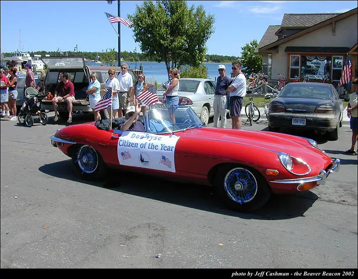 A decorated red vintage convertible passes spectators beside the waterfront during a parade.