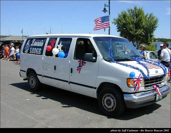 A white van decorated with patriotic streamers, balloons, bows, and flags is surrounded by spectators near the waterfront.
