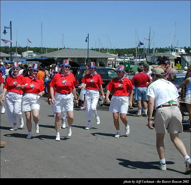 Women in matching red shirts and white shorts march past a marina during a parade.