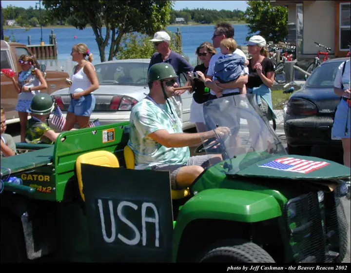 A green decorated utility vehicle with a driver passes spectators beside the water.