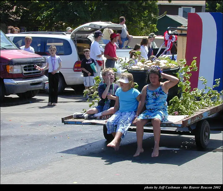 Children ride on a plant-filled parade trailer beside parked vehicles and a large red, white, and blue decoration.