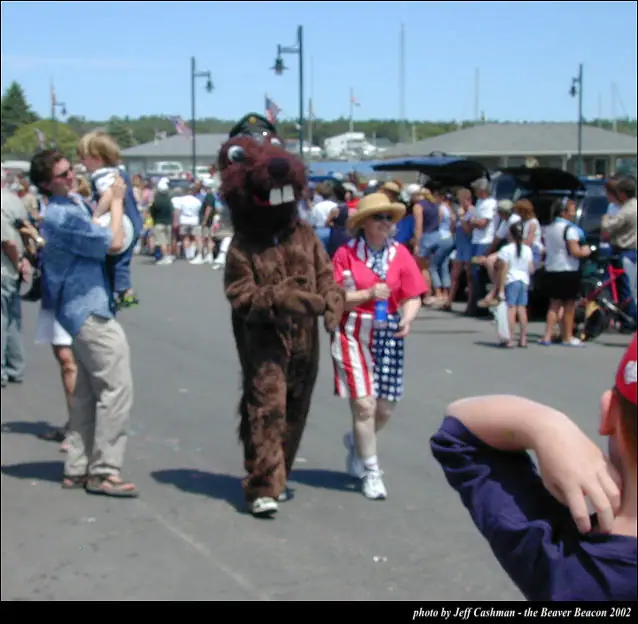 A beaver mascot and costumed marcher move through a crowded outdoor parade while spectators watch.