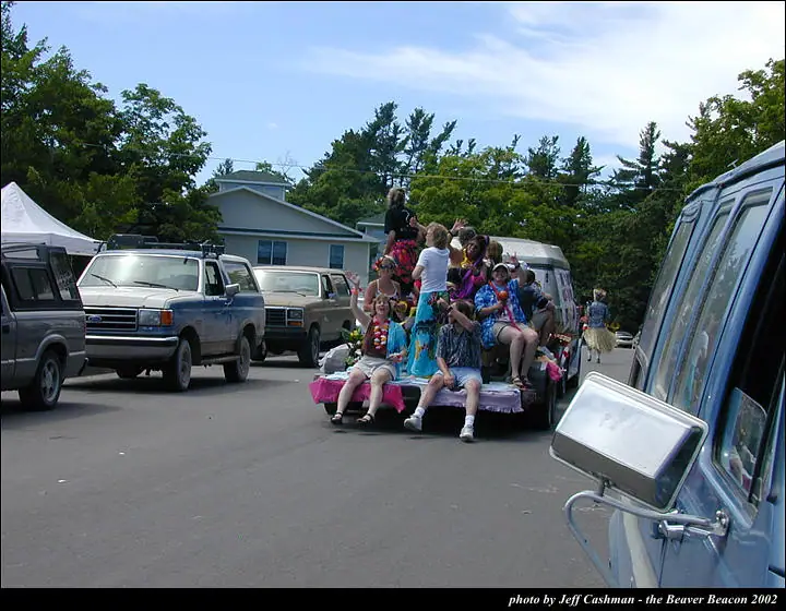 A decorated trailer carries people in colorful costumes among parked vehicles on a tree-lined street.