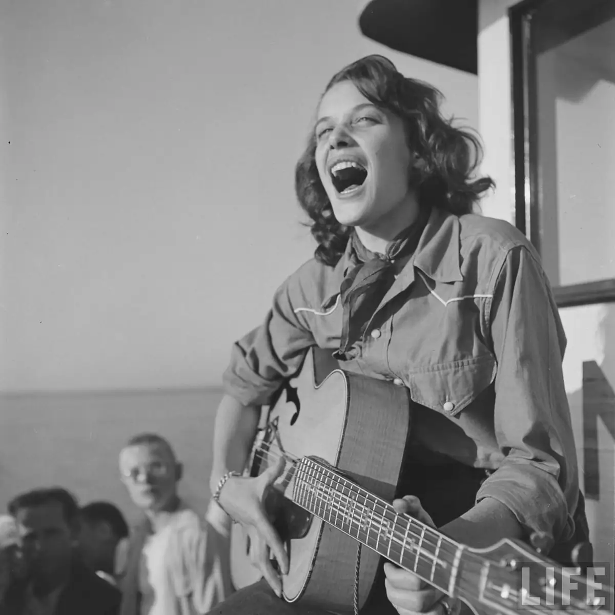 Black-and-white photograph of a singer playing an acoustic guitar before an audience.