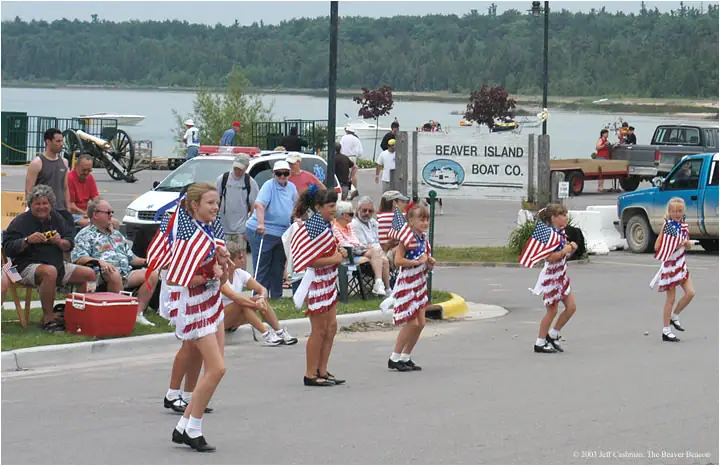 Five girls in patriotic outfits march past spectators along a waterfront road.