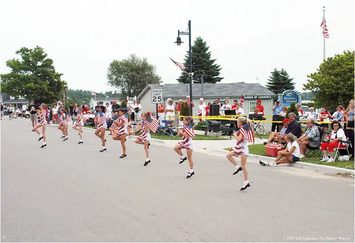 Young marchers in patriotic outfits carry flags along a street as spectators watch from the sidewalks.