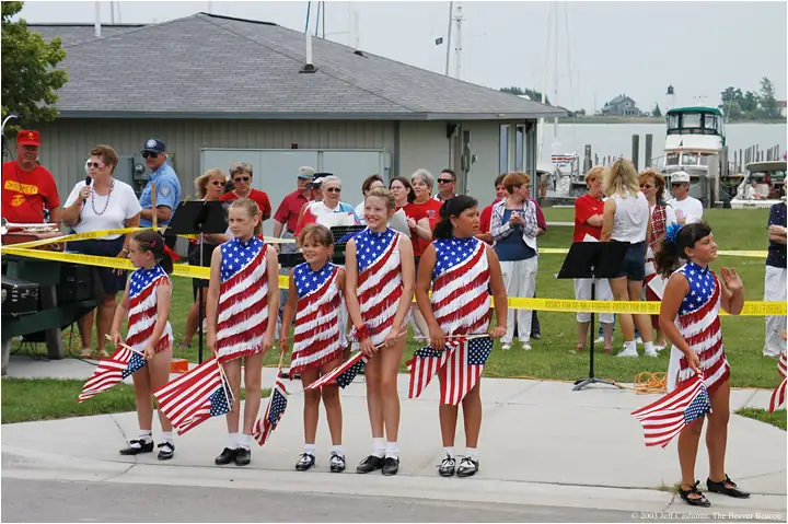 Children in patriotic costumes hold American flags during an outdoor waterfront parade as spectators watch.