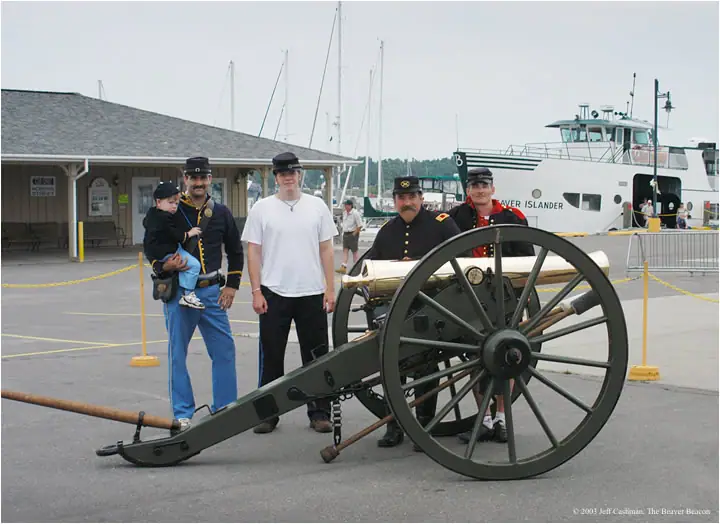 Five people, including uniformed adults and a child, pose beside a wheeled cannon near a ferry.