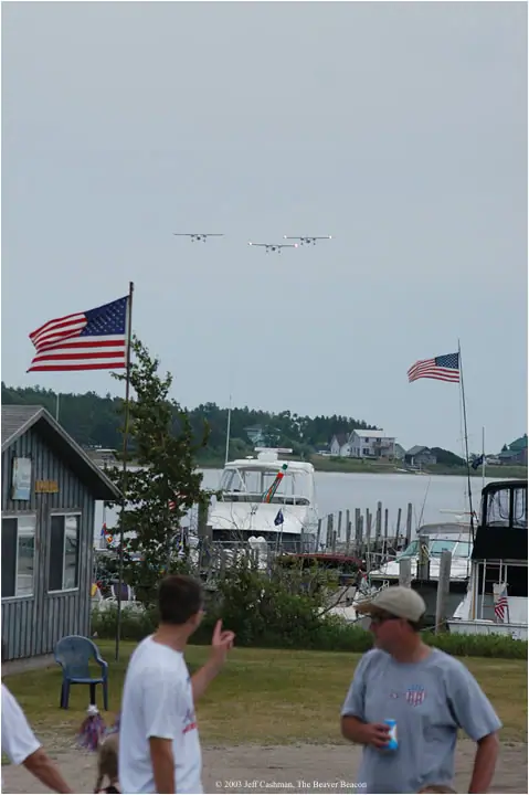 People watch aircraft flying over a waterfront harbor with boats, docks, houses, and large American flags.