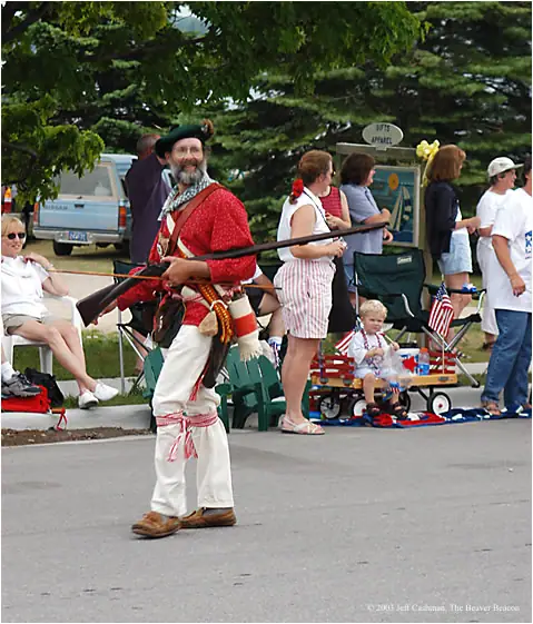 A costumed historical reenactor carrying a rifle walks past spectators and a child in a decorated wagon.