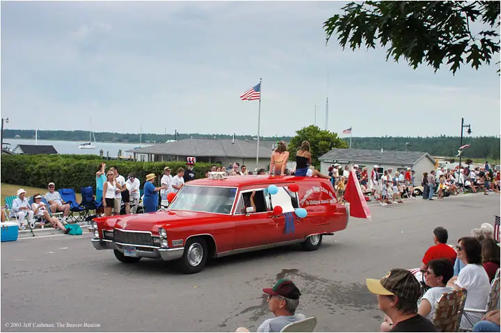 A decorated red hearse carries two people through a crowded parade beside the waterfront.