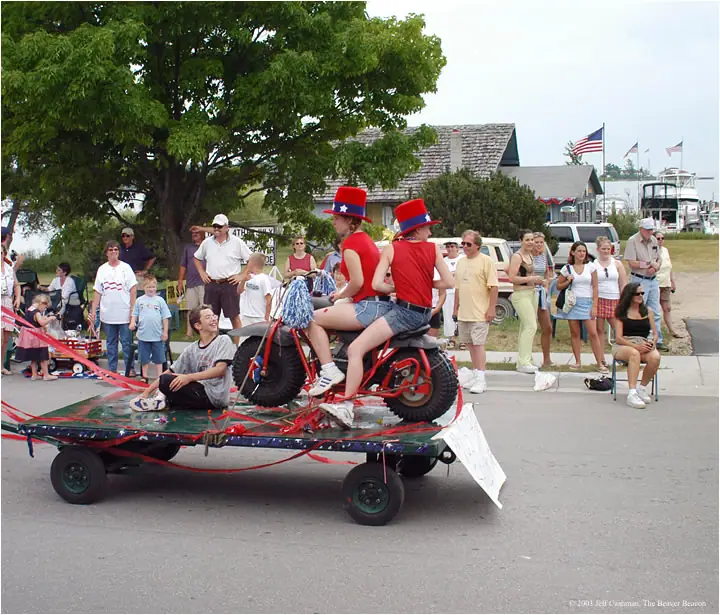 A decorated parade float carries a red motorcycle and costumed riders past a crowd and waterfront buildings.