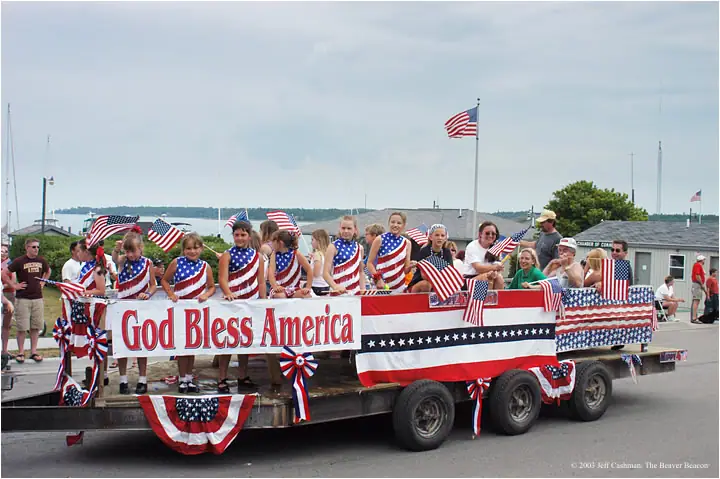 Children and adults ride a patriotic parade float decorated with American flags and bunting.