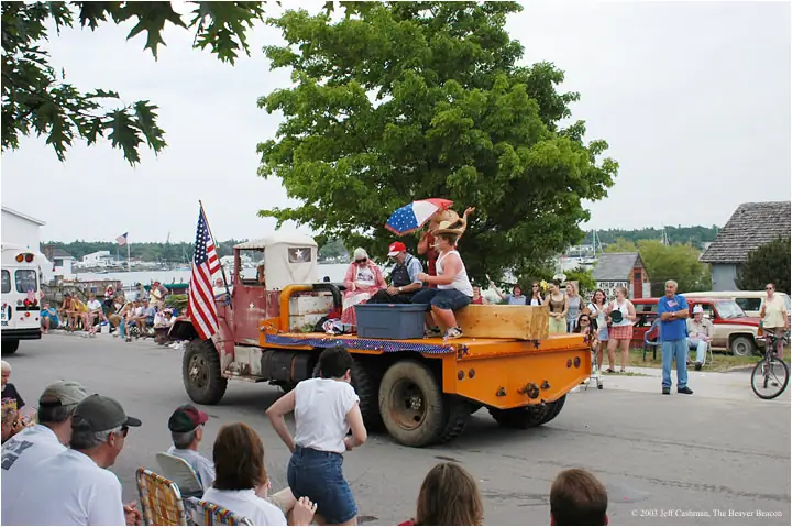 A decorated orange truck carrying people passes seated spectators along a waterfront street.