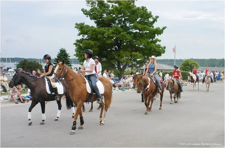 Horseback riders proceed past seated spectators during a waterfront parade.