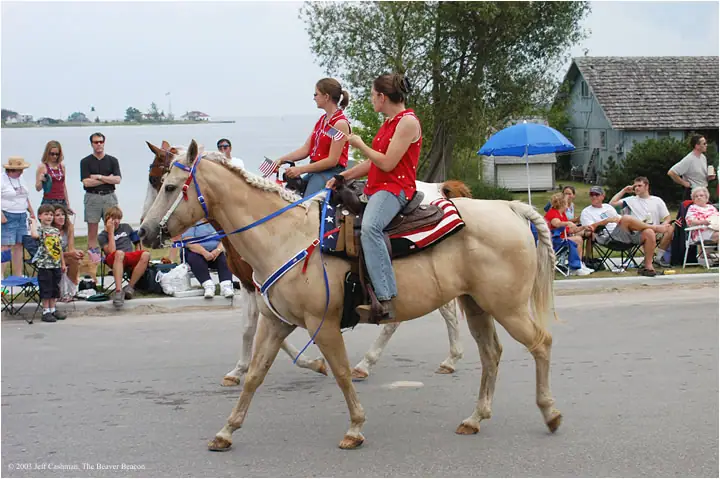 Two riders on decorated horses pass spectators along a waterfront street.