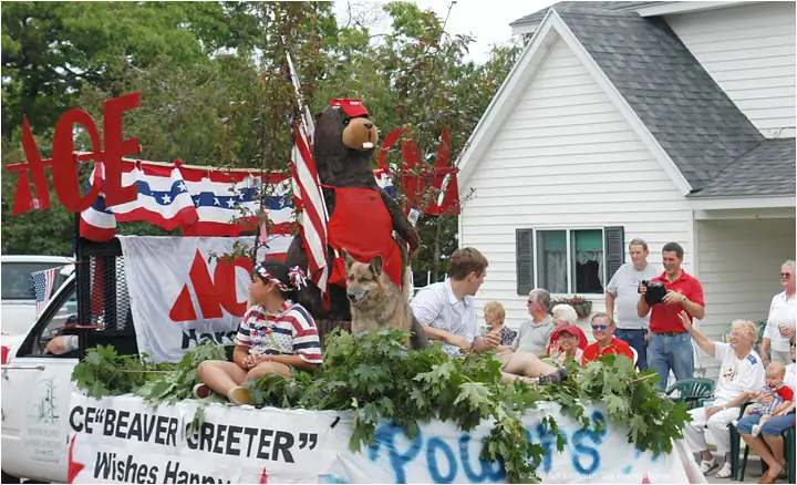 A parade float carries a beaver mascot, a dog, patriotic decorations, and a seated child past spectators.