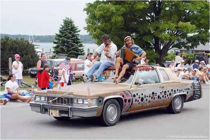 Decorated sedan carries two musicians playing violin and accordion during a parade.
