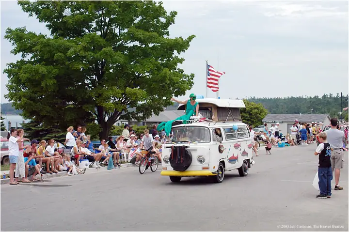 Decorated white van leads a parade past seated spectators, with a person on its roof and an American flag overhead.