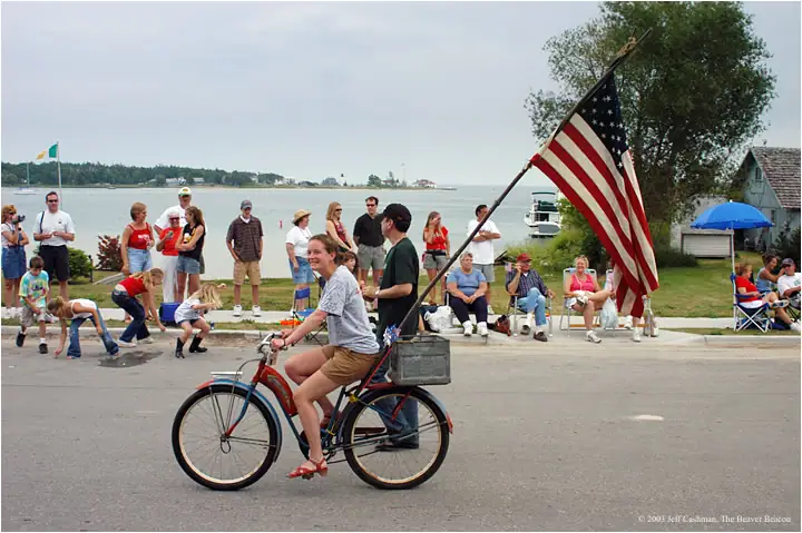 A woman pedals a bicycle carrying a second rider and a large American flag past seated spectators.