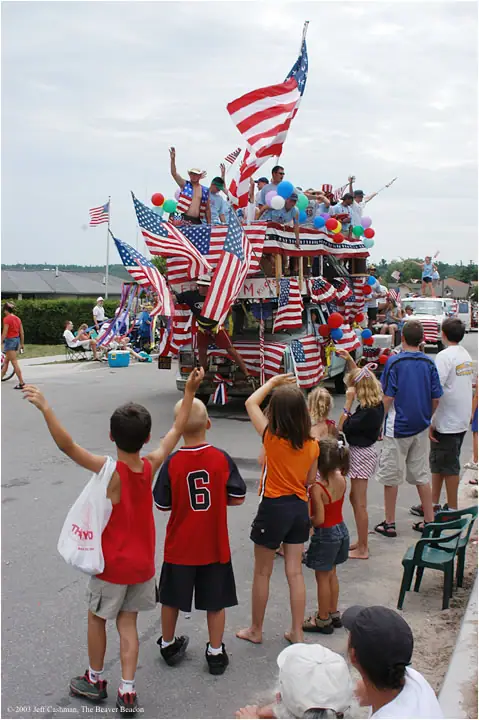 A flag-covered parade truck carries waving people past children and spectators lining the street.