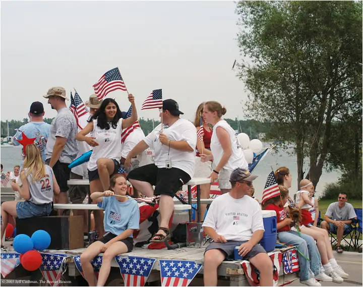 People ride a flag-decorated parade float beside the water while holding flags and balloons.