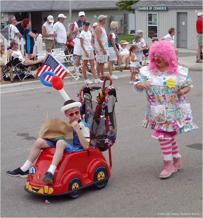 A child rides a red pedal car in a parade while a brightly costumed clown walks alongside.