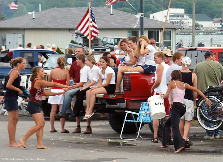A crowd gathers around a red pickup truck beside a waterfront, with flags, bicycles, and buildings behind them.