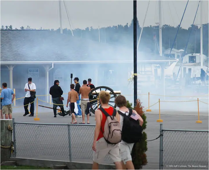 A cannon demonstration takes place beside a marina, with smoke, spectators, and sailboat masts in the background.