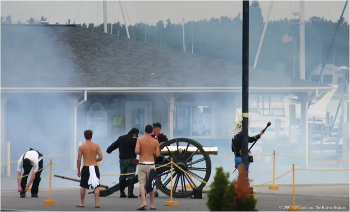 People gather around a large cannon amid smoke outside a waterfront building, with boats and a flag behind them.