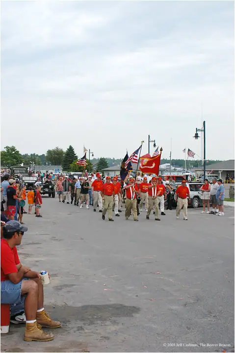 A uniformed parade group carries American and colorful flags past spectators near a waterfront.