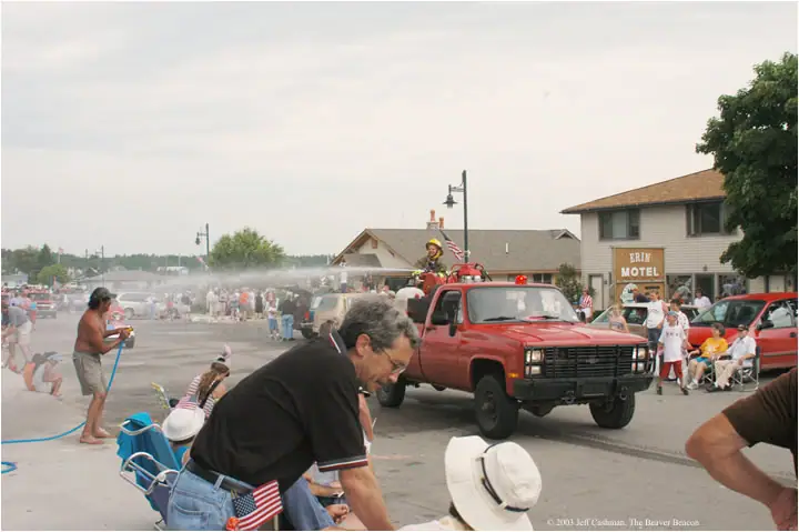 A red pickup sprays water along a street as a uniformed person and spectators watch.