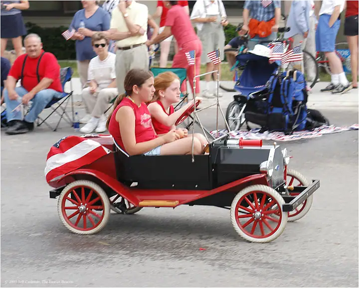 Two girls ride in a small flag-decorated vintage car during a parade.