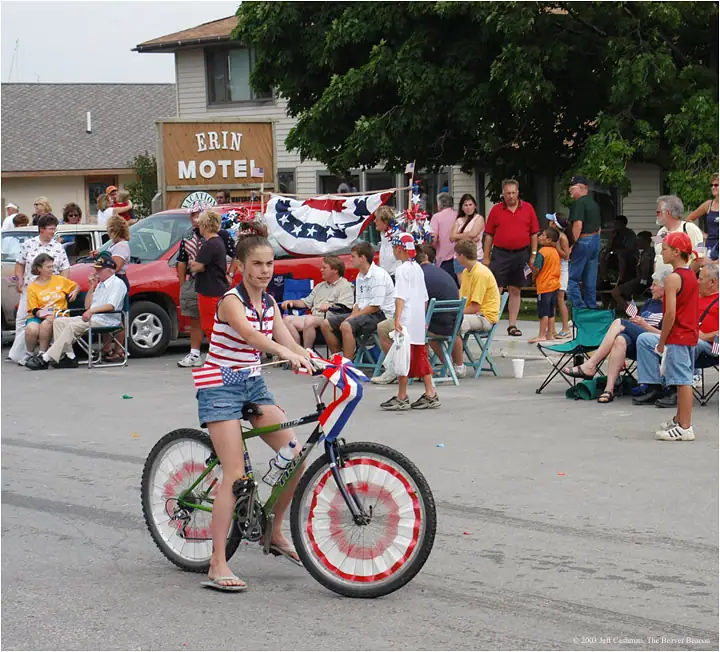 A girl rides a red, white, and blue decorated bicycle past seated spectators on a street.